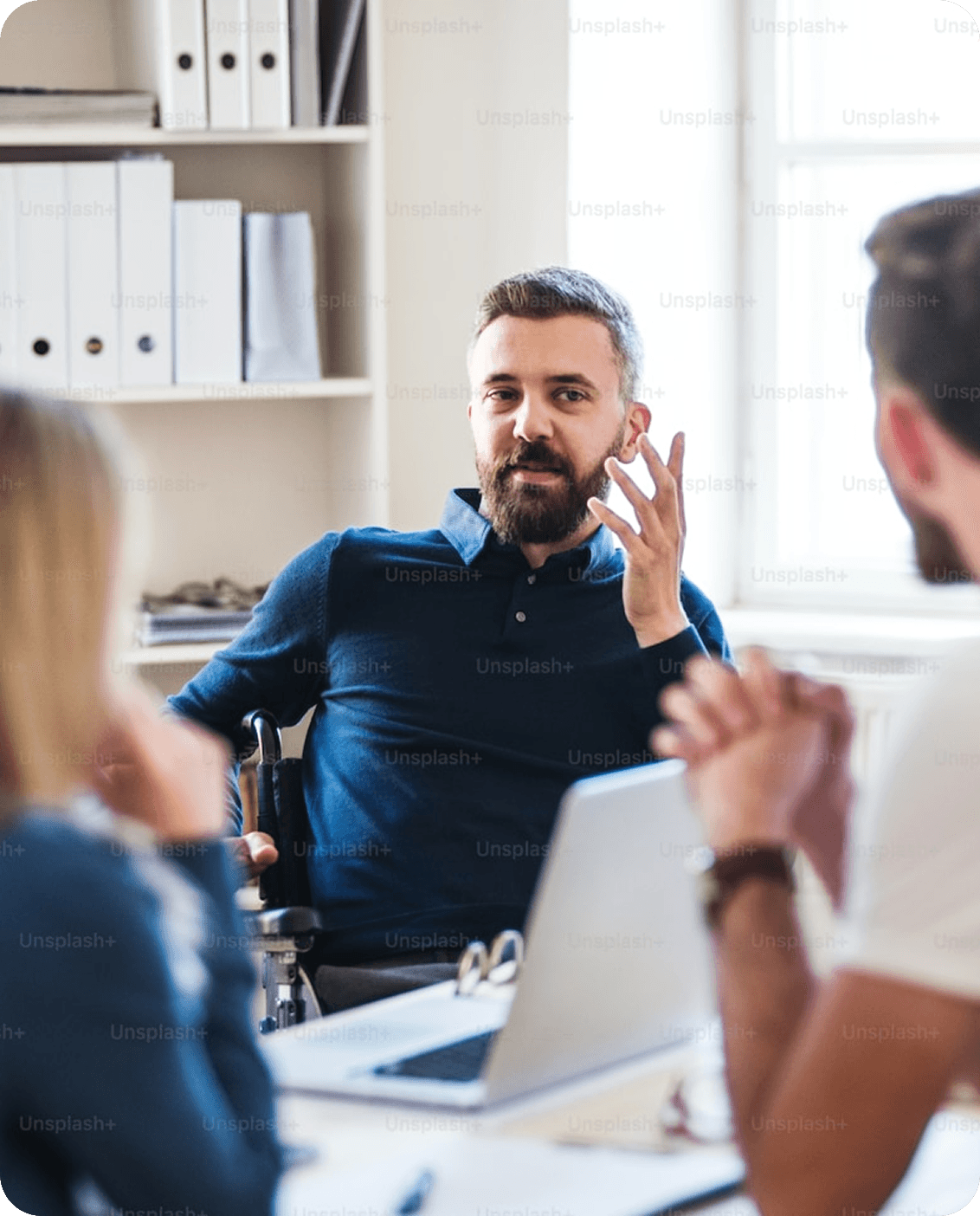 A professional man with a beard sitting in a wheelchair and gesturing while leading a meeting with colleagues in a bright, modern office.