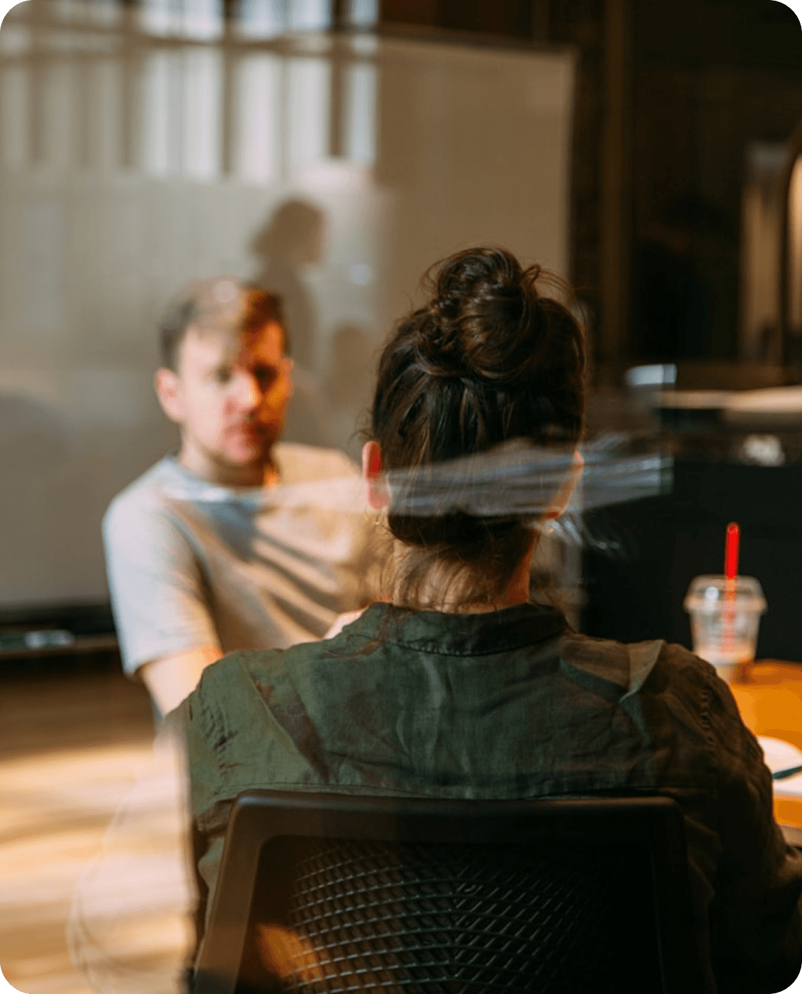 Colleagues in a warm, modern office: one teammate seen from behind in a green shirt at a wooden table, facing a teammate in soft focus across a collaborative conversation with natural light and glass partitions in the background.