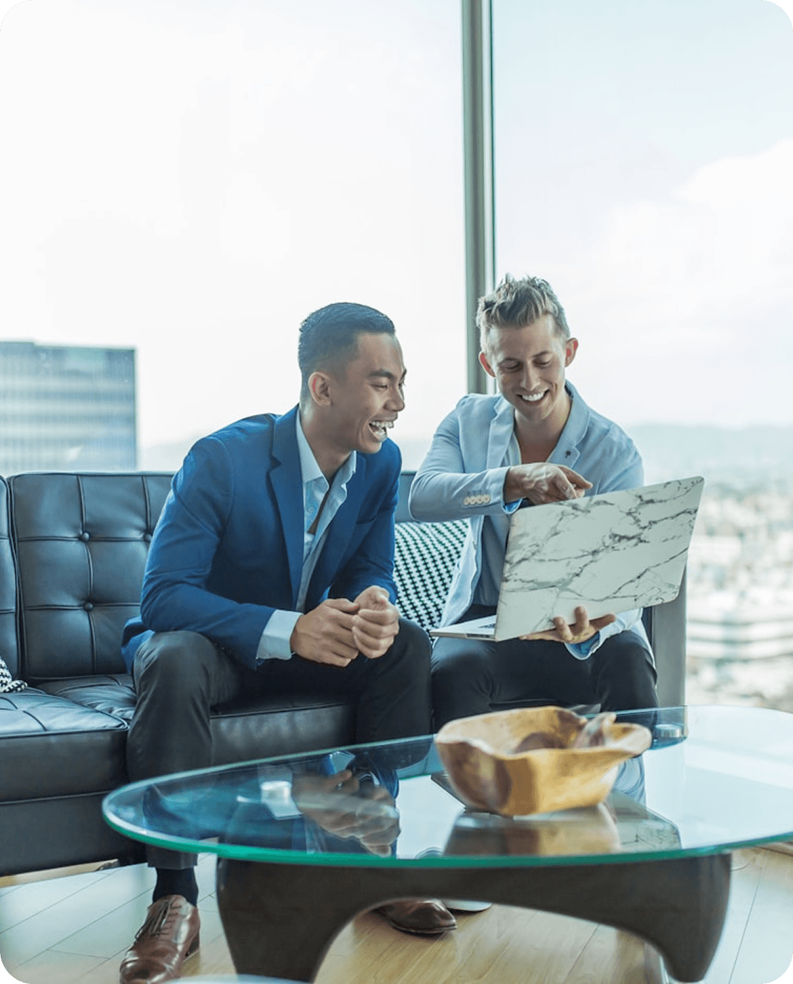 Two professional colleagues laughing and collaborating over a laptop in a modern, sunlit high-rise office with a city view through floor-to-ceiling windows.